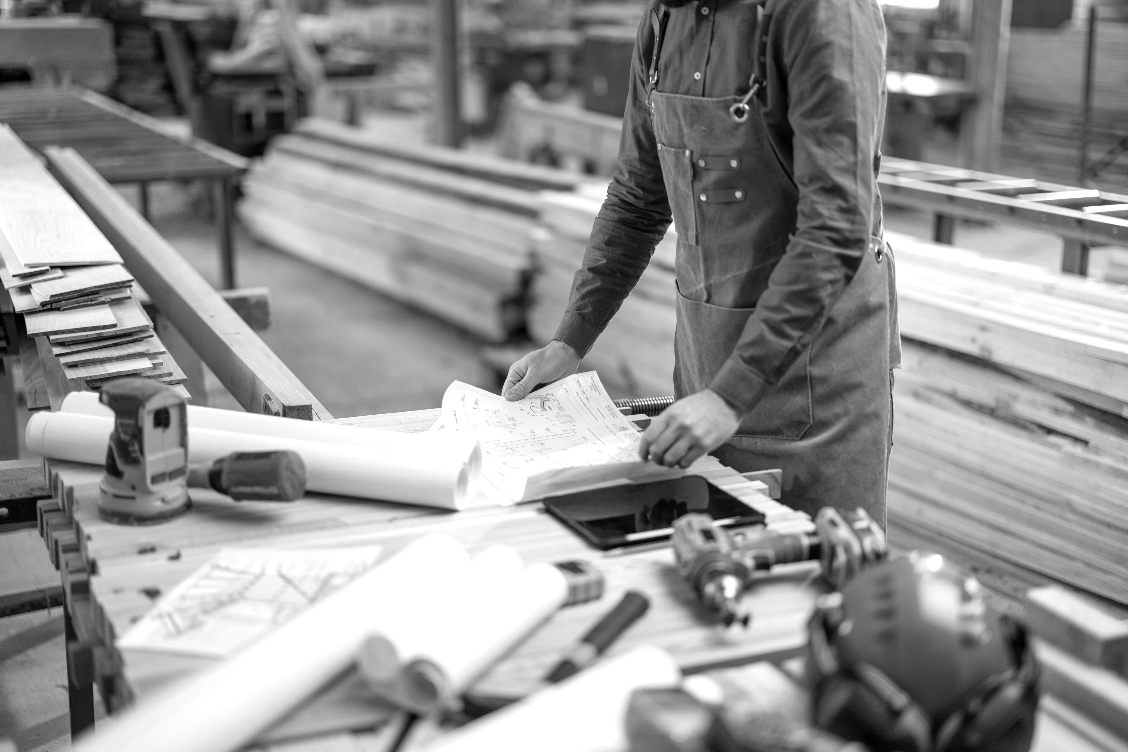 Woodworker in an apron reviews blueprints on a workbench with tools.